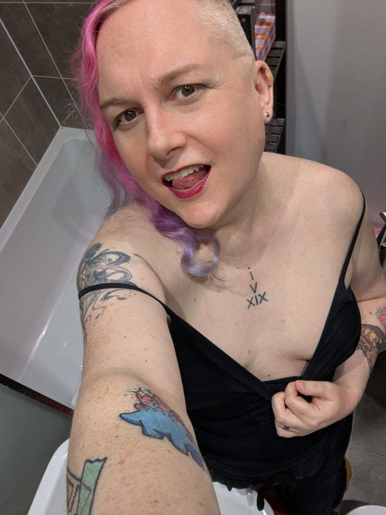 Selfie of me in the bathroom, pulling my black strappy top down so you can see some boob, and making eye contact with the camera.