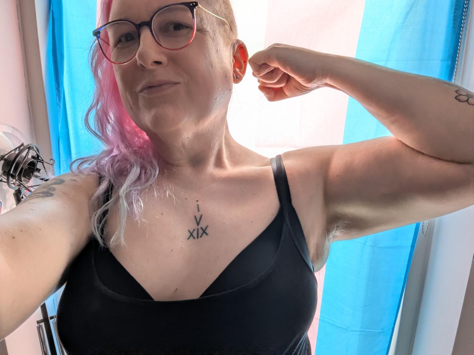 Selfie of me flexing my free arm in front of a backlit trans flag